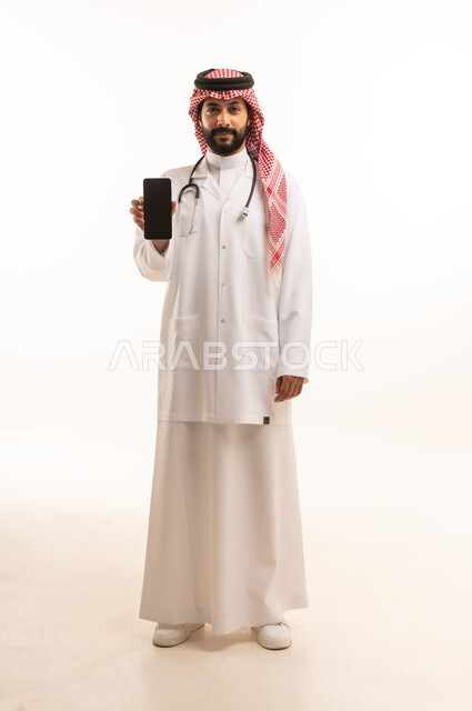 عرض شاشة سوداء فارغة عبر الهاتف المحمول، مفهوم الطب والرعاية الصحية، بورتريه لطبيب عربي خليجي سعودي يرتدي الثوب التقليدي مع المعطف والسماعة الطبية يقدم الخدمات الصحية للمرضى، مهنة ووظيفة طبية، العمل في القطاع الصحي في المملكة، الجسم بالطول الكامل، خلفيه بيضاء