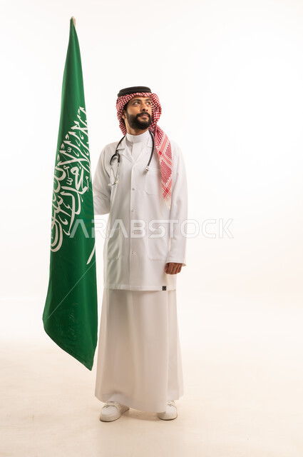 إحياء ذكرى اليوم الوطني السعودي 23 سبتمبر، مفهوم الطب والرعاية الصحية، بورتريه لطبيب عربي خليجي سعودي يرتدي الثوب التقليدي مع المعطف والسماعة الطبية يقدم الخدمات الصحية للمرضى ويحمل بيده علم المملكة، العمل في القطاع الصحي في المملكة، الجسم بالطول الكامل، خلفيه بيضاء