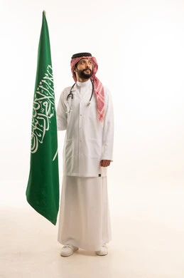 إحياء ذكرى اليوم الوطني السعودي 23 سبتمبر، مفهوم الطب والرعاية الصحية، بورتريه لطبيب عربي خليجي سعودي يرتدي الثوب التقليدي مع المعطف والسماعة الطبية يقدم الخدمات الصحية للمرضى ويحمل بيده علم المملكة، العمل في القطاع الصحي في المملكة، الجسم بالطول الكامل، خلفيه بيضاء