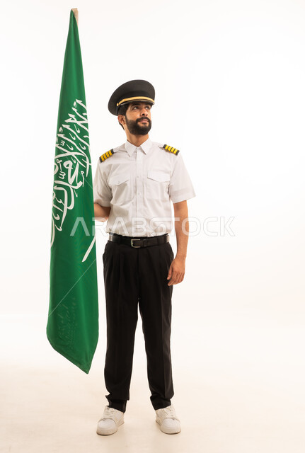 الاحتفال بذكرى اليوم الوطني السعودي 23 سبتمبر, مفهوم السياحة والسفر، طاقم الاستقبال والترحيب، العمل في مجال الطيران، رحلات جوية سياحية, خطوط الطيران السعودية, بورتريه لكابتن طيارعربي سعودي خليجي يرتدي الزي يحمل بيده علم المملكة، الجسم بالطول الكامل، خلفيه بيضاء