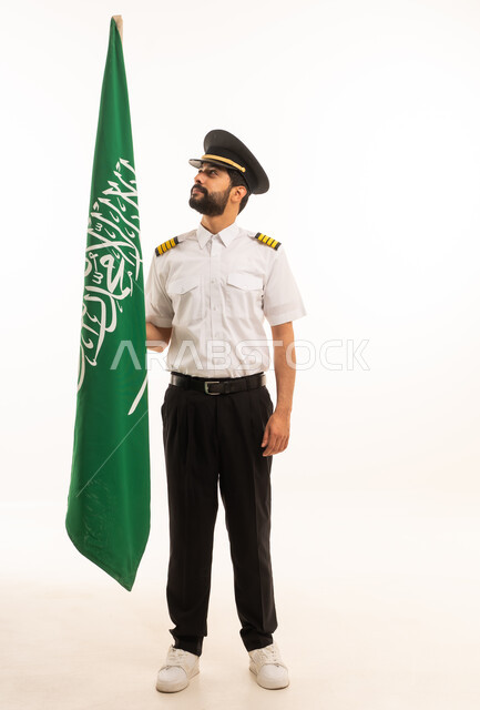 الاحتفال بذكرى اليوم الوطني السعودي 23 سبتمبر, مفهوم السياحة والسفر، طاقم الاستقبال والترحيب، العمل في مجال الطيران، رحلات جوية سياحية, خطوط الطيران السعودية, بورتريه لكابتن طيارعربي سعودي خليجي يرتدي الزي يحمل بيده علم المملكة، الجسم بالطول الكامل، خلفيه بيضاء