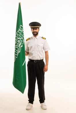 Celebrating Saudi National Day on September 23, the concept of tourism and travel, reception and welcome staff, working in the aviation field, tourist flights, Saudi Airlines, portrait of a Saudi Arabian Gulf Arab pilot wearing a uniform holding the flag of the Kingdom, full body, white background
