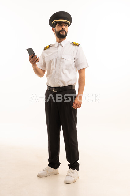 استخدام الهاتف المحمول، مفهوم السياحة والسفر، طاقم الاستقبال والترحيب بالركاب، العمل في مجال الطيران، رحلات جوية سياحية, خطوط الطيران السعودية, بورتريه لكابتن طيار عربي سعودي خليجي مبتسم يرتدي الزي الرسمي يقف باستقامة، الجسم بالطول الكامل، خلفيه بيضاء