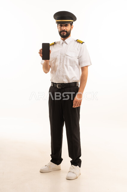 عرض شاشة سوداء فارغة عبر الهاتف المحمول، طاقم الاستقبال والترحيب بالركاب، العمل في مجال الطيران، رحلات جوية سياحية,  مفهوم السياحة والسفر، خطوط الطيران السعودية, بورتريه لكابتن طيار عربي سعودي خليجي يرتدي الزي الرسمي يقف باستقامة، الجسم بالطول الكامل، خلفيه بيضاء