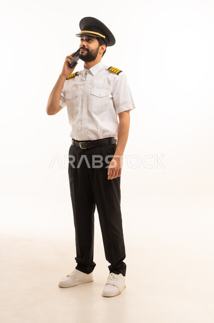 اجراء مكالمة هاتفية عبر الهاتف المحمول، طاقم الاستقبال والترحيب بالركاب، العمل في مجال الطيران، رحلات جوية سياحية,  مفهوم السياحة والسفر، خطوط الطيران السعودية, بورتريه لكابتن طيار عربي سعودي خليجي يرتدي الزي الرسمي يقف باستقامة، الجسم بالطول الكامل، خلفيه بيضاء