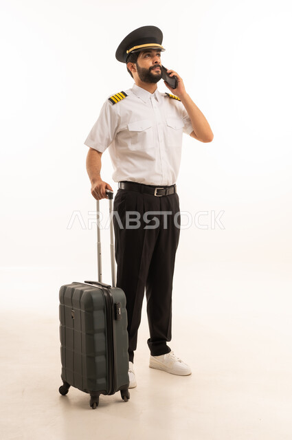 اجراء مكالمة هاتفية عبر الهاتف المحمول، طاقم الاستقبال والترحيب بالركاب، العمل في مجال الطيران، رحلات جوية سياحية,  مفهوم السياحة والسفر، خطوط الطيران السعودية, بورتريه لكابتن طيار عربي سعودي خليجي يرتدي الزي الرسمي ويجر الحقيبة، الجسم بالطول الكامل، خلفيه بيضاء
