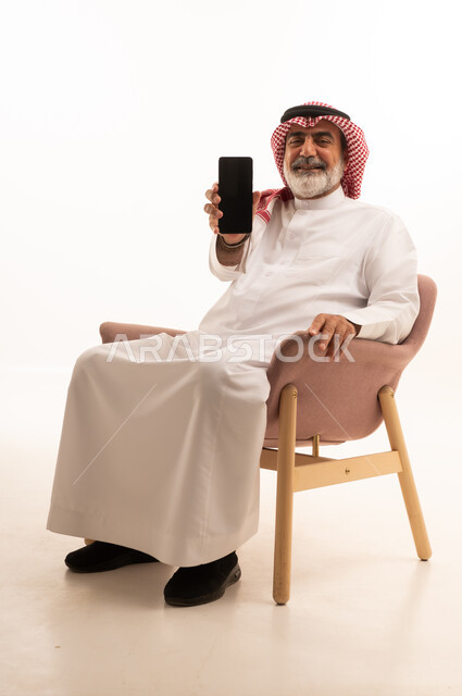 عرض شاشة سوداء فارغة عبر الجوال، بورتريه لرجل عربي خليجي سعودي يرتدي الشماغ والثوب التقليدي يمسك بيده الهاتف المحمول، الدردشة والتواصل مع الأهل و الأصدقاء، استخدام جهاز تقني حديث ومتطور، العمل عن بعد وانجاز المهام المطلوبة، خلفية بيضاء