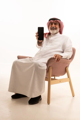 عرض شاشة سوداء فارغة عبر الجوال، بورتريه لرجل عربي خليجي سعودي يرتدي الشماغ والثوب التقليدي يمسك بيده الهاتف المحمول، الدردشة والتواصل مع الأهل و الأصدقاء، استخدام جهاز تقني حديث ومتطور، العمل عن بعد وانجاز المهام المطلوبة، خلفية بيضاء