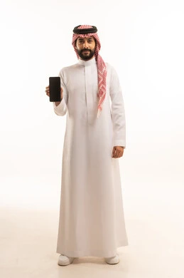 عرض شاشة سوداء فارغة عبر الجوال، بورتريه لرجل عربي خليجي سعودي يرتدي الشماغ والثوب التقليدي يمسك بيده الهاتف المحمول، الدردشة والتواصل مع الأهل و الأصدقاء، استخدام جهاز تقني حديث ومتطور، العمل عن بعد وانجاز المهام المطلوبة، الجسم بالطول الكامل، خلفية بيضاء