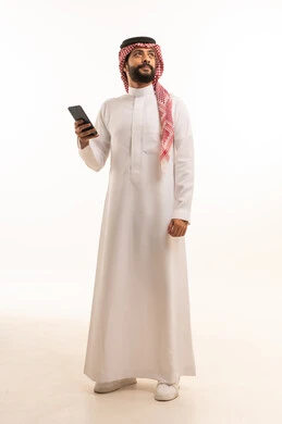 استخدام جهاز تقني حديث ومتطور، العمل عن بعد وانجاز المهام المطلوبة، تصفح مواقع التواصل الاجتماعي، بورتريه لرجل عربي خليجي سعودي يرتدي الشماغ والثوب التقليدي يمسك بيده الهاتف المحمول، الدردشة والتواصل مع الأهل و الأصدقاء، الجسم بالطول الكامل، خلفية بيضاء