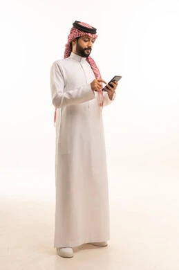 استخدام جهاز تقني حديث ومتطور، العمل عن بعد وانجاز المهام المطلوبة، تصفح مواقع التواصل الاجتماعي، بورتريه لرجل عربي خليجي سعودي يرتدي الشماغ والثوب التقليدي يمسك بيده الهاتف المحمول، الدردشة والتواصل مع الأهل و الأصدقاء، الجسم بالطول الكامل، خلفية بيضاء
