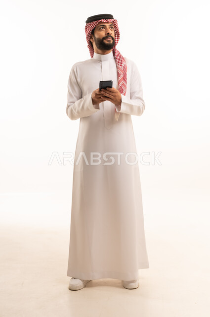 استخدام جهاز تقني حديث ومتطور، العمل عن بعد وانجاز المهام المطلوبة، تصفح مواقع التواصل الاجتماعي، بورتريه لرجل عربي خليجي سعودي يرتدي الشماغ والثوب التقليدي يمسك بيده الهاتف المحمول، الدردشة والتواصل مع الأهل و الأصدقاء، الجسم بالطول الكامل، خلفية بيضاء