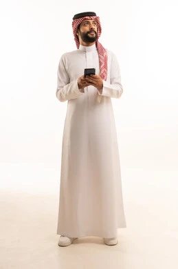 استخدام جهاز تقني حديث ومتطور، العمل عن بعد وانجاز المهام المطلوبة، تصفح مواقع التواصل الاجتماعي، بورتريه لرجل عربي خليجي سعودي يرتدي الشماغ والثوب التقليدي يمسك بيده الهاتف المحمول، الدردشة والتواصل مع الأهل و الأصدقاء، الجسم بالطول الكامل، خلفية بيضاء