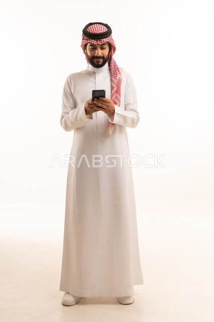استخدام جهاز تقني حديث ومتطور، العمل عن بعد وانجاز المهام المطلوبة، تصفح مواقع التواصل الاجتماعي، بورتريه لرجل عربي خليجي سعودي يرتدي الشماغ والثوب التقليدي يمسك بيده الهاتف المحمول، الدردشة والتواصل مع الأهل و الأصدقاء، الجسم بالطول الكامل، خلفية بيضاء