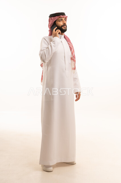 اجراء مكالمة هاتفية عبر الجوال، استخدام جهاز تقني حديث ومتطور، العمل عن بعد وانجاز المهام المطلوبة، بورتريه لرجل عربي خليجي سعودي يرتدي الشماغ والثوب التقليدي يمسك بيده الهاتف المحمول، الدردشة والتواصل مع الأهل و الأصدقاء، الجسم بالطول الكامل، خلفية بيضاء