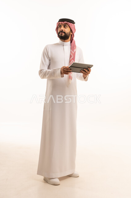 Working remotely and completing required tasks, managing and organizing business affairs through technical devices, an administrative profession and job, integrating work with technology, a portrait of a Saudi Arabian Gulf man wearing a traditional thobe and shemagh working on a tablet, using a modern and advanced technical device, a white background