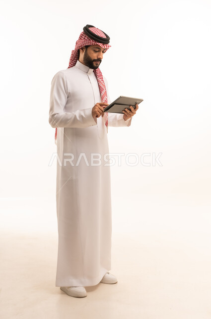 Working remotely and completing required tasks, managing and organizing business affairs through technical devices, an administrative profession and job, integrating work with technology, a portrait of a Saudi Arabian Gulf man wearing a traditional thobe and shemagh working on a tablet, using a modern and advanced technical device, a white background
