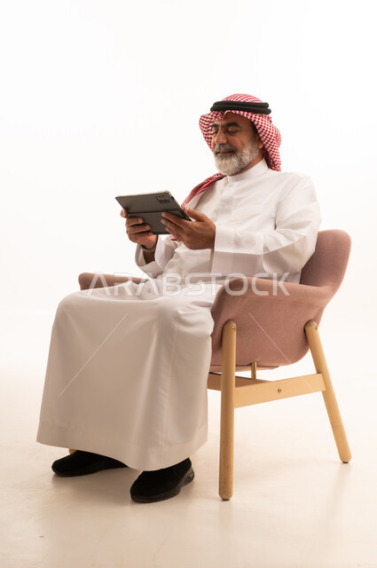 العمل عن بعد وانجاز المهام المطلوبة، إدارة شؤون الأعمال وتنظيمها عبر الاجهزة التقنية، مهنة ووظيفة ادارية، دمج العمل مع التكنولوجيا، بورتريه لرجل عربي خليجي سعودي يرتدي الثوب التقليدي والشماغ يعمل على الجهاز اللوحي، استخدام جهاز تقني حديث ومتطور، خلفية بيضاء 