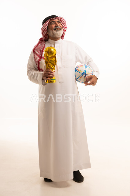 تعابير الفرح والسرور والحماس للمنافسة، مفهوم الربح والفوز، تشجيع الفريق السعودي، التتويج بالبطولة والحصول على المركز الأول، بورتريه لرجل عربي خليجي سعودي يرتدي الثوب التقليدي والشماغ يحمل بيده كأس كرة قدم ذهبي, صورة الجسم بالطول الكامل, خلفية بيضاء