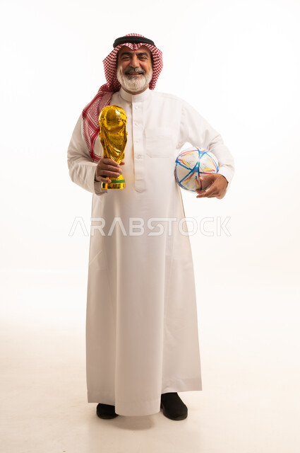تعابير الفرح والسرور والحماس للمنافسة، مفهوم الربح والفوز، تشجيع الفريق السعودي، التتويج بالبطولة والحصول على المركز الأول، بورتريه لرجل عربي خليجي سعودي يرتدي الثوب التقليدي والشماغ يحمل بيده كأس كرة قدم ذهبي, صورة الجسم بالطول الكامل, خلفية بيضاء