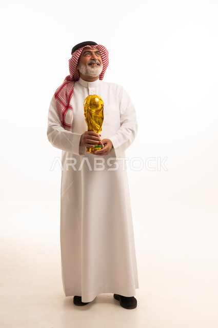 تعابير الفرح والسرور والحماس للمنافسة، مفهوم الربح والفوز، تشجيع الفريق السعودي، التتويج بالبطولة والحصول على المركز الأول، بورتريه لرجل عربي خليجي سعودي يرتدي الثوب التقليدي والشماغ يحمل بيده كأس كرة قدم ذهبي, صورة الجسم بالطول الكامل, خلفية بيضاء