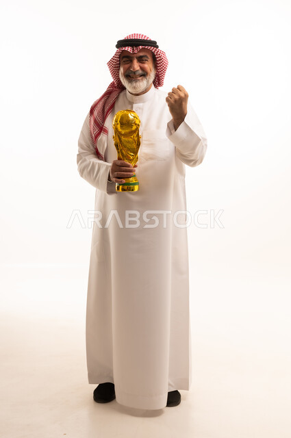 تعابير الفرح والسرور والحماس للمنافسة، مفهوم الربح والفوز، تشجيع الفريق السعودي، التتويج بالبطولة والحصول على المركز الأول، بورتريه لرجل عربي خليجي سعودي يرتدي الثوب التقليدي والشماغ يحمل بيده كأس كرة قدم ذهبي, صورة الجسم بالطول الكامل, خلفية بيضاء