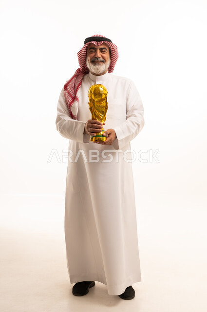 تعابير الفرح والسرور والحماس للمنافسة، مفهوم الربح والفوز، تشجيع الفريق السعودي، التتويج بالبطولة والحصول على المركز الأول، بورتريه لرجل عربي خليجي سعودي يرتدي الثوب التقليدي والشماغ يحمل بيده كأس كرة قدم ذهبي, صورة الجسم بالطول الكامل, خلفية بيضاء