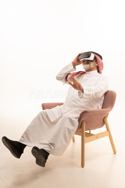 التفاعل والاندماج ومحاكاة العالم الخيالي، بورتريه لرجل عربي خليجي سعودي مبتسم يرتدي الثوب التقليدي والشماغ بجلس على كرسي مريح يضع نظارة الواقع الافتراضي يرفع يده لمحاولة لمس شيء ما، المتعة والتسلية بالمشاهدة عبر نظارات ثلاثية الابعاد، خلفية بيضاء