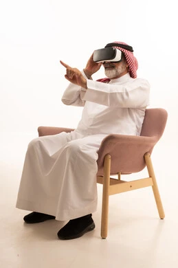 التفاعل والاندماج ومحاكاة العالم الخيالي، بورتريه لرجل عربي خليجي سعودي مبتسم يرتدي الثوب التقليدي والشماغ بجلس على كرسي مريح يضع نظارة الواقع الافتراضي يرفع يده لمحاولة لمس شيء ما، المتعة والتسلية بالمشاهدة عبر نظارات ثلاثية الابعاد، خلفية بيضاء