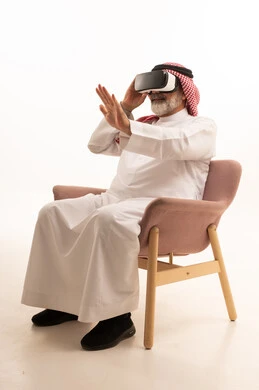 التفاعل والاندماج ومحاكاة العالم الخيالي، بورتريه لرجل عربي خليجي سعودي مبتسم يرتدي الثوب التقليدي والشماغ بجلس على كرسي مريح يضع نظارة الواقع الافتراضي يرفع يده لمحاولة لمس شيء ما، المتعة والتسلية بالمشاهدة عبر نظارات ثلاثية الابعاد، خلفية بيضاء