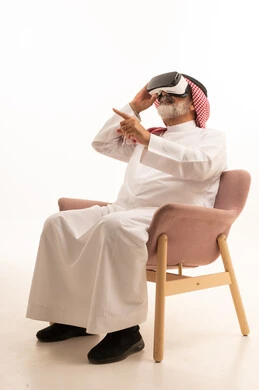 Interaction, integration and simulation of the imaginary world, portrait of a smiling Saudi Arabian Gulf man wearing the traditional thobe and shemagh sitting on a comfortable chair wearing virtual reality glasses and raising his hand to try to touch something, enjoyment and entertainment by watching through 3D glasses, white background