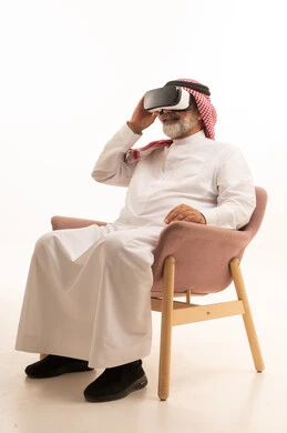التفاعل والاندماج ومحاكاة العالم الخيالي، بورتريه لرجل عربي خليجي سعودي مبتسم يرتدي الثوب التقليدي والشماغ بجلس على كرسي مريح يضع نظارة الواقع الافتراضي يرفع يده لمحاولة لمس شيء ما، المتعة والتسلية بالمشاهدة عبر نظارات ثلاثية الابعاد، خلفية بيضاء