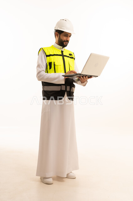 استخدام جهاز تقني حديث ومتطور، مشاريع إعمارية سعودية وطنية، مفهوم الهندسة المعمارية، الاهتمام بالتطور العمراني بسواعد المهندسين السعوديين, بورتريه لمهندس عربي خليجي سعودي يرتدي سترة العمل وخوذة الحماية يحمل بيده الحاسوب المحمول، الجسم بالطول الكامل، خلفية بيضاء