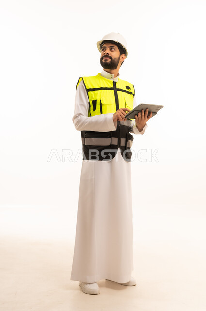 مفهوم الهندسة المعمارية، الاهتمام بالتطور العمراني بسواعد المهندسين السعوديين, استخدام جهاز تقني حديث ومتطور، مشاريع إعمارية سعودية وطنية، بورتريه لمهندس عربي خليجي سعودي يرتدي سترة العمل وخوذة الحماية يحمل بيده الجهاز اللوحي، الجسم بالطول الكامل، خلفية بيضاء
