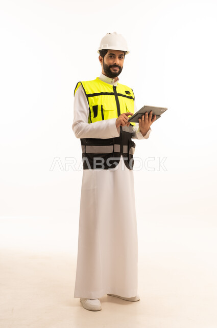 مفهوم الهندسة المعمارية، الاهتمام بالتطور العمراني بسواعد المهندسين السعوديين, استخدام جهاز تقني حديث ومتطور، مشاريع إعمارية سعودية وطنية، بورتريه لمهندس عربي خليجي سعودي يرتدي سترة العمل وخوذة الحماية يحمل بيده الجهاز اللوحي، الجسم بالطول الكامل، خلفية بيضاء