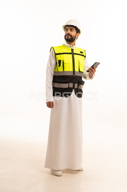 دمج التكنولوجيا والتقنية الحديثة في مجال الهندسة المعمارية، الاهتمام بالتطور العمراني بسواعد المهندسين السعوديين، مشاريع إعمارية سعودية وطنية، بورتريه لمهندس عربي خليجي سعودي يرتدي سترة العمل وخوذة الحماية يحمل بيده الهاتف المحمول، الجسم بالطول الكامل، خلفية بيضاء