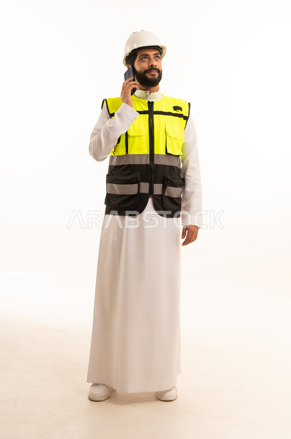 دمج التكنولوجيا والتقنية الحديثة في مجال الهندسة المعمارية، الاهتمام بالتطور العمراني بسواعد المهندسين السعوديين، مشاريع إعمارية سعودية وطنية، بورتريه لمهندس عربي خليجي سعودي يرتدي سترة العمل وخوذة الحماية يحمل بيده الهاتف المحمول، الجسم بالطول الكامل، خلفية بيضاء