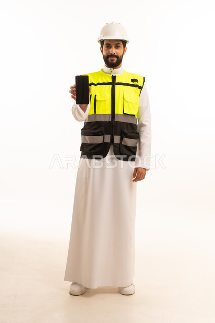 دمج التكنولوجيا والتقنية الحديثة في مجال الهندسة المعمارية، الاهتمام بالتطور العمراني بسواعد المهندسين السعوديين، مشاريع إعمارية سعودية وطنية، بورتريه لمهندس عربي خليجي سعودي يرتدي سترة العمل وخوذة الحماية يحمل بيده الهاتف المحمول، الجسم بالطول الكامل، خلفية بيضاء