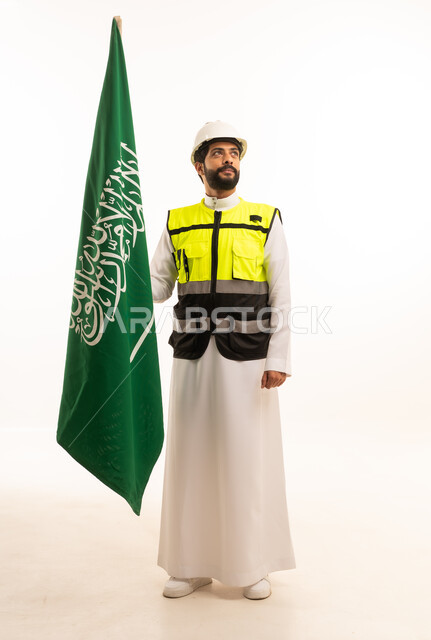 الاحتفال بذكرى اليوم الوطني السعودي 23 سبتمبر، مفهوم الهندسة المعمارية، الاهتمام بالتطور العمراني بسواعد المهندسين السعوديين، مشاريع إعمارية وطنية، بورتريه لمهندس عربي خليجي سعودي يرتدي سترة العمل وخوذة الحماية يقف بجانب علم المملكة، الجسم بالطول الكامل، خلفية بيضاء