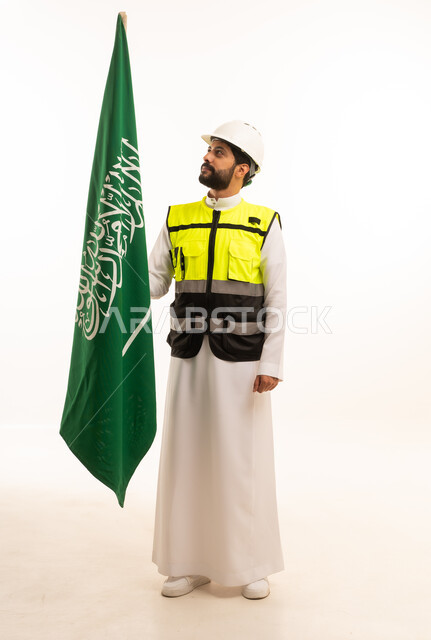 الاحتفال بذكرى اليوم الوطني السعودي 23 سبتمبر، مفهوم الهندسة المعمارية، الاهتمام بالتطور العمراني بسواعد المهندسين السعوديين، مشاريع إعمارية وطنية، بورتريه لمهندس عربي خليجي سعودي يرتدي سترة العمل وخوذة الحماية يقف بجانب علم المملكة، الجسم بالطول الكامل، خلفية بيضاء