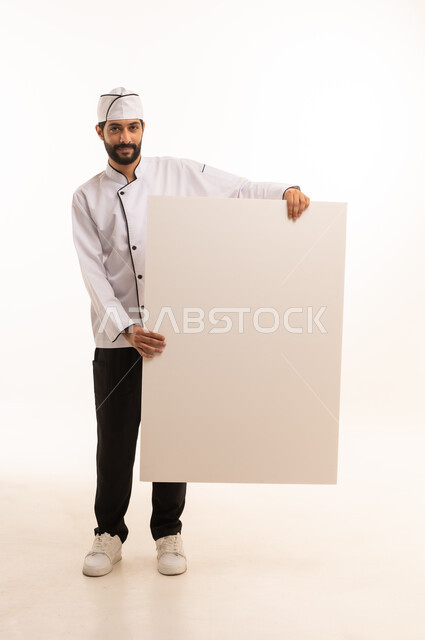 موك اب ابيض كبير, احتراف مهنة طهي الطعام السعودي, الوقوف باستقامة وتعابير السعادة, بورتريه لشيف عربي خليجي سعودي يرتدي يونيفورم الطبخ يمسك بيده لوحة بيضاء فارغة، الجسم بالطول الكامل, خلفية بيضاء