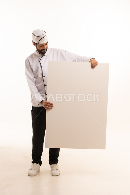 موك اب ابيض كبير, احتراف مهنة طهي الطعام السعودي, الوقوف باستقامة وتعابير السعادة, بورتريه لشيف عربي خليجي سعودي يرتدي يونيفورم الطبخ يمسك بيده لوحة بيضاء فارغة، الجسم بالطول الكامل, خلفية بيضاء