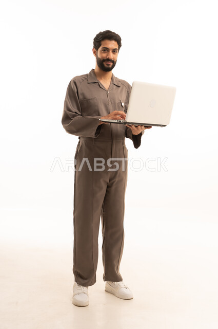 استخدام جهاز تقني حديث, المهارة و اتقان العمل، استعمال معدات الصيانة لاجراء التصليحات، بورتريه لرجل عربي سعودي خليجي يرتدي يونيفورم العمل يحمل الحاسب المحمول, الجسم بالطول الكامل، خلفية بيضاء