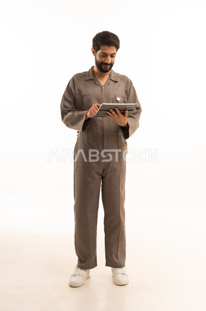 استخدام جهاز تقني حديث, المهارة و اتقان العمل، استعمال معدات الصيانة لاجراء التصليحات، بورتريه لرجل عربي سعودي خليجي يرتدي يونيفورم العمل يحمل جهاز التابلت اللوحي, الجسم بالطول الكامل، خلفية بيضاء