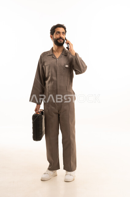 Making a voice call, skill and mastery of work, using maintenance equipment to make repairs, portrait of a Saudi Arabian Gulf man wearing a work uniform holding a tool bag and mobile phone, full body, white background