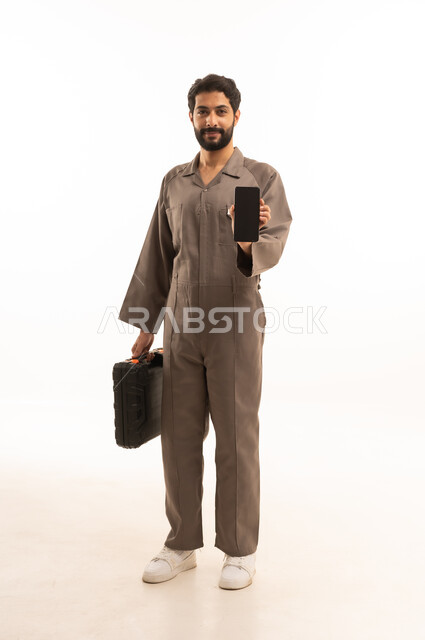 Blank black screen, skill and workmanship, using maintenance equipment to make repairs, portrait of a Saudi Arabian Gulf man wearing work uniform holding a tool bag and mobile phone, full body, white background