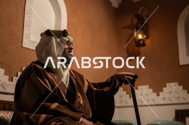 الحكمة والتجربة الطويلة في الحياة، صورة جانبية لرجل عربي سعودي مسن يرتدي الزي العربي التقليدي يجلس على كرسي خشبي داخل غرفة تراثية بديكورات جدران مزخرفة، يرتدي عباءة واسعة وعقالًا على رأسه، ويبدو بلحية بيضاء، بيده عكاز يستند إليها .