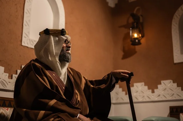 الحكمة والتجربة الطويلة في الحياة، صورة جانبية لرجل عربي سعودي مسن يرتدي الزي العربي التقليدي يجلس على كرسي خشبي داخل غرفة تراثية بديكورات جدران مزخرفة، يرتدي عباءة واسعة وعقالًا على رأسه، ويبدو بلحية بيضاء، بيده عكاز يستند إليها .