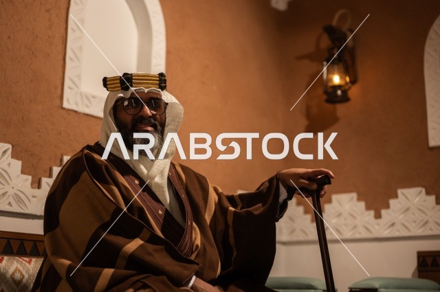 صورة جانبية لرجل عربي سعودي مسن يرتدي الزي العربي التقليدي يجلس على كرسي خشبي داخل غرفة تراثية بديكورات جدران مزخرفة، يرتدي عباءة واسعة وعقالًا على رأسه، ويبدو بلحية بيضاء، بيده عكاز يستند إليها، الحكمة والتجربة الطويلة في الحياة.