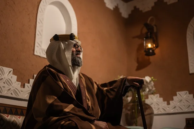 الأصالة والحنين للأيام القديمة، صورة جانبية عربي سعودي لرجل مسن يرتدي الزي العربي التقليدي يجلس على كرسي خشبي داخل غرفة تراثية بديكورات جدران مزخرفة، يرتدي عباءة واسعة وعقالًا على رأسه، ويبدو بلحية بيضاء، بيده عكاز يستند إليها، الحكمة والتجربة الطويلة في الحياة.